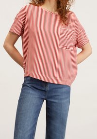 Kvinna som bär en röd och vit vertikalt randig kortärmad skjorta med ficka, ihop med blå jeans, stående mot en enfärgad bakgrund.