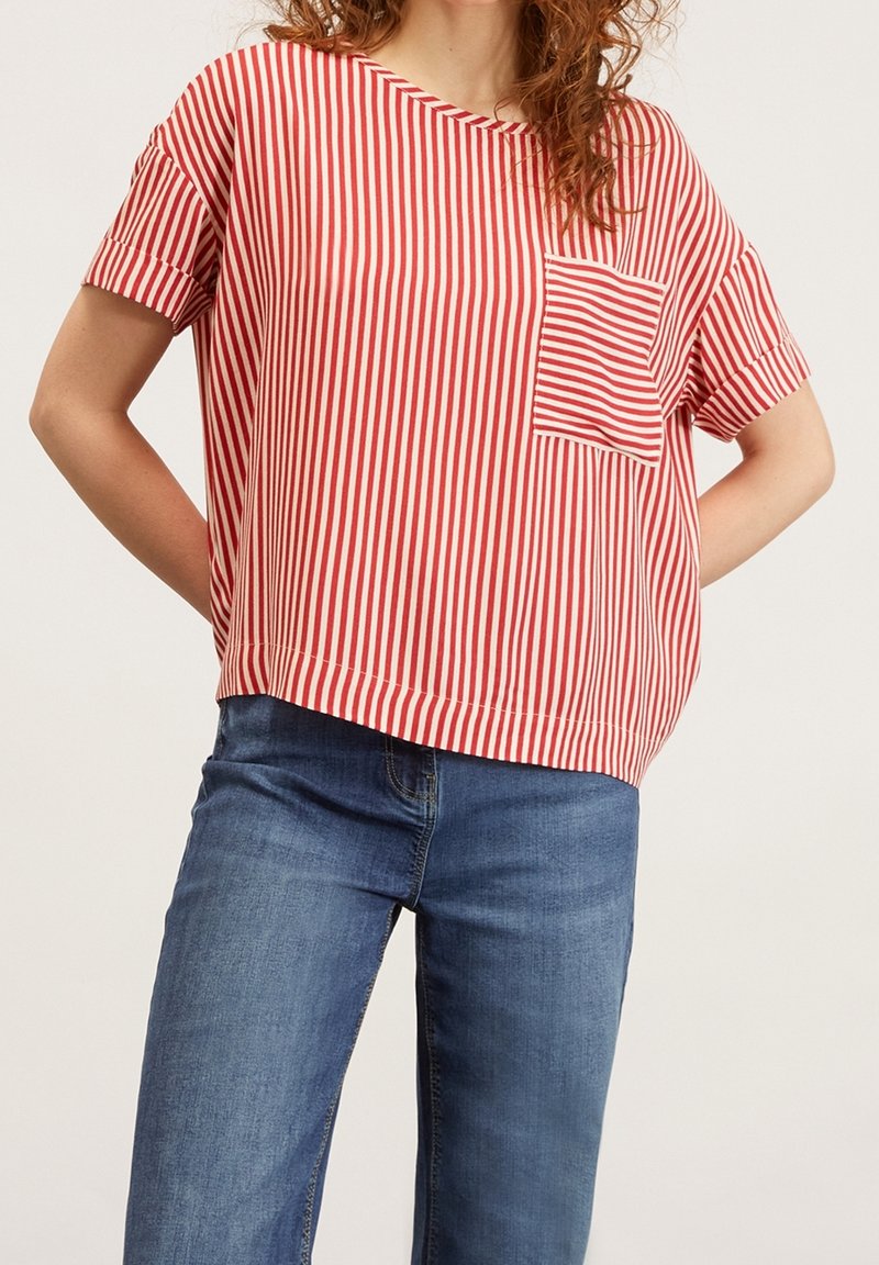 Kvinna som bär en röd och vit vertikalt randig kortärmad skjorta med ficka, ihop med blå jeans, stående mot en enfärgad bakgrund.