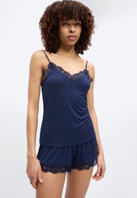 Junge Frau mit lockigem Haar, die ein marineblaues Camisole- und Shorts-Set mit weißen Punkten und Spitzenbesatz trägt, steht vor einem schlichten Hintergrund.