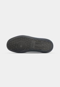 Semelle en caoutchouc noir avec des motifs de traction circulaires et hexagonaux, présentant un bord lisse et un détail de logo imprimé près du talon.