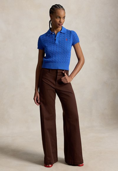 Femme aux cheveux tressés portant un polo en maille bleu manches courtes et un pantalon marron à jambes larges, debout avec une main sur la hanche.