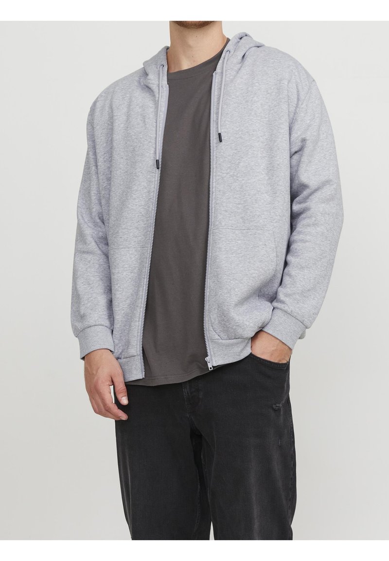 Homme portant un sweat à capuche gris clair zippé sur un t-shirt gris foncé avec un pantalon noir, une main dans la poche, cadré au niveau du cou et au milieu de la cuisse.