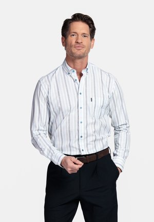 Man met een wit, lichtblauw en beige gestreept overhemd met knopen, donkere broek en een bruine riem, staand met één hand in zijn zak.