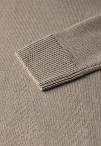 Beige knitted sweater with ribbed cuff and textured fabric. Close-up view highlights the soft, fine-knit material and detailed stitching.