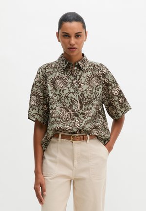 Frau trägt ein kurzärmeliges, grün-braun geblümtes Hemd, das in beige Hosen mit braunem Gürtel gesteckt ist.