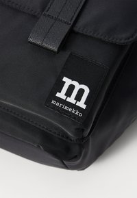Borsa nera in nylon con chiusura a pattina. Presenta una tracolla testurizzata e un'etichetta logo rettangolare con lettering bianco. Superficie liscia.