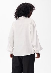 Biała bluzka z długimi rękawami o gładkiej fakturze, z kołnierzykiem i bufkowymi rękawami, noszona na czarnych spodniach.