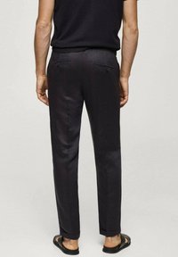 Dark, tailored pants with a smooth texture, two back pockets, and rolled cuffs. Worn with black flip-flops, viewed from the back.