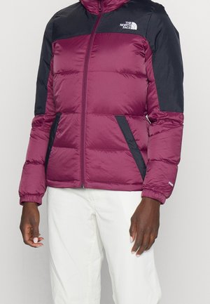 Osoba ubrana w bordowo-czarną pikowaną kurtkę North Face z zamkiem z przodu oraz białe spodnie, stojąca na jednolitym tle.