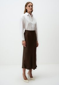 Camicia bianca con bottoni e gonna midi in satin marrone, con orlo svasato e texture liscia. Modella che indossa décolleté a punta in avorio.
