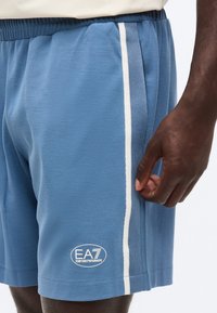 Μπλε αθλητικά σορτς με λευκές ρίγες στο πλάι και το λογότυπο Emporio Armani EA7 στο αριστερό μηρό, φορεμένα από άτομο με σκούρο δέρμα ορατό.