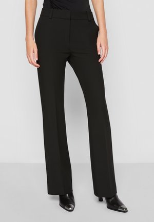 Femme portant un pantalon noir taille haute sur mesure et des chaussures noires à bouts pointus avec embouts métalliques, debout devant un fond gris uni.