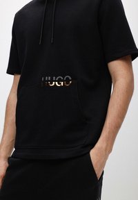 Sort kortærmet hættebluse med en frontlomme. Har et metallic sølv- og rosaguld "HUGO" logo. Lavet af blødt stof.