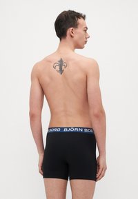 Schwarze Boxer-Briefs mit einem blauen Logo-Bund. Der Stoff wirkt glatt und passt gut, zeigt ein einfaches Design ohne Muster.
