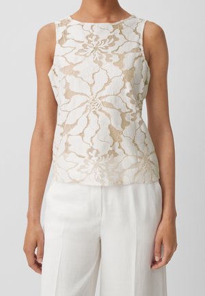 Ærmeløs beige bluse i blonder med blomstermønster, båret med løstsiddende hvide bukser med højtaljet snit.