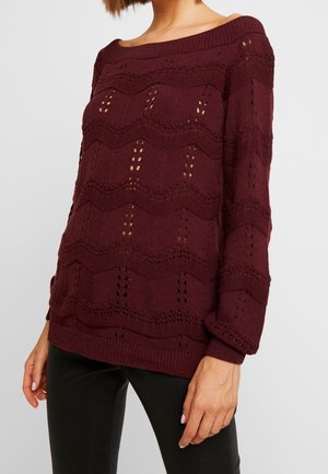 Maglione a maglia bordeaux con motivo ondulato e dettagli traforati. Presenta un scollo off-shoulder e polsini a coste. Abbinato a pantaloni neri.
