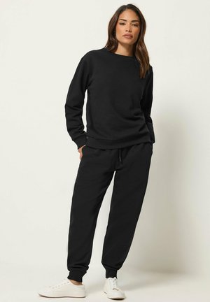 Donna in piedi con le mani in tasca, indossa una felpa nera, pantaloni da jogging neri e scarpe da ginnastica bianche su uno sfondo bianco uniforme.