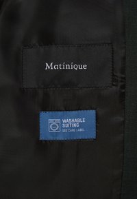 Étiquette en tissu noire avec texte blanc "Matinique" au-dessus d'une étiquette bleue indiquant "Washable Suiting See Care Label" avec une icône de machine à laver.