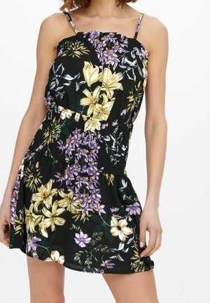 Kobieta w czarnej, bezrękawnikowej sukience w kwiaty w kolorze żółtym i fioletowym, z cienkimi ramiączkami i dopasowaną talią.