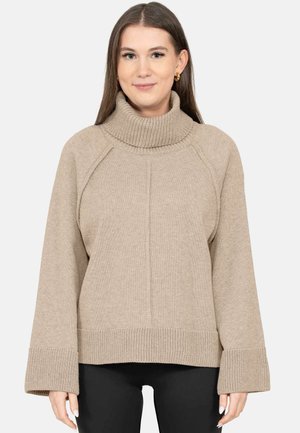 Jonge vrouw met een beige gebreide coltrui met brede boorden en een zwarte broek, staand tegen een effen achtergrond.