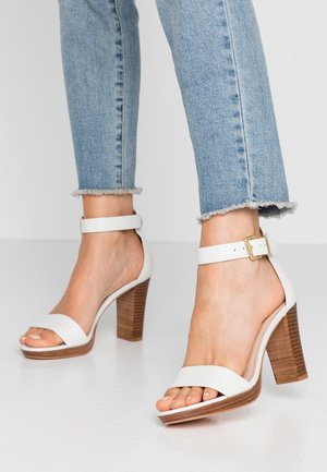 Białe sandały na obcasie z szerokim paskiem na palcach, z klamrą przy kostce i drewnianym słupkiem. Noszone z dżinsami o postrzępionych nogawkach.