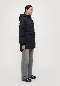 Svart boblejakke med hette, med glidelås foran, to store lommer, og et teksturert materiale. Kombinert med lyseblå vide jeans og svarte joggesko.
