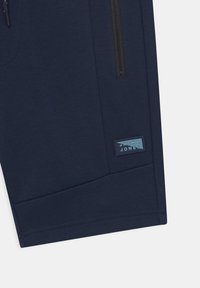 Mørkeblå joggebukser med glidelåslomme, flate sømmer, og et rektangulært logo-patch i lys blå med preget tekst.