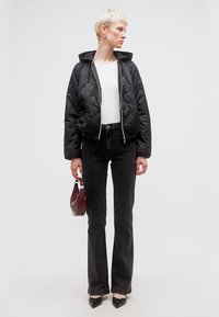 Γυναίκα με κοντά ξανθά μαλλιά, φορούσα μαύρο καπιτονέ μπουφάν, λευκή μπλούζα, μαύρα τζιν, μαύρα μυτερά παπούτσια, κρατώντας μια μικρή σκούρα κόκκινη τσάντα.