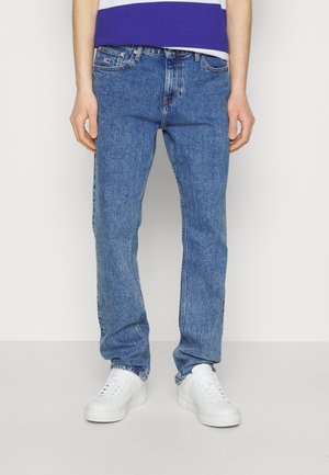 Homme portant un jean denim bleu et des baskets blanches, debout devant un fond uni.