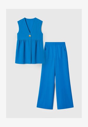 Kék ujjatlan V-nyakú gombos felső, széles szárú nadrággal, oldalán zsebekkel, fehér háttéren bemutatva.