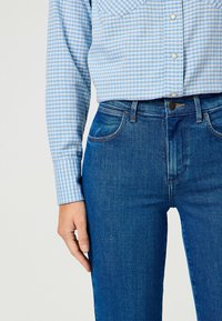 Camisa de cuadros azules con cuello, combinada con unos jeans de color azul oscuro. La camisa tiene botones y mangas largas; los jeans cuentan con un diseño clásico de cinco bolsillos.