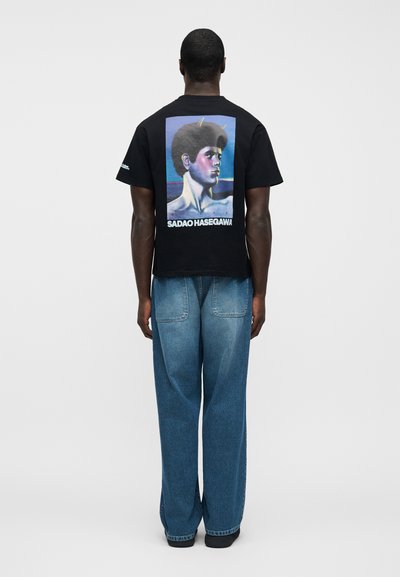 Muž otočený chrbtom má na sebe čierne tričko s farebným portrétom a nápisom "SADAO HASEGAWA" na chrbte, doplnené o voľné modré džínsy a čierne topánky.