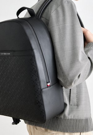 Personne portant un sac à dos noir Tommy Hilfiger par-dessus une petite veste grise à carreaux et un pantalon beige.