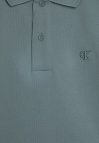 Polo tričko v svetlom modrom materiáli, s textúrovaným pletením, dvomi gombíkmi a malým vyšitým logom "CK" na hrudi.