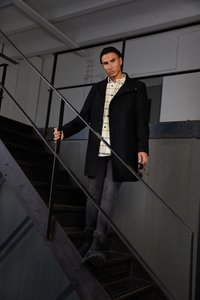 Casaco preto com gola alta, sobre uma camisa xadrez. Calças cinzas e botas pretas. Escadas com corrimãos de metal em um ambiente industrial.