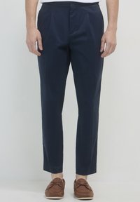 Homme portant un pantalon ajusté bleu marine et des mocassins en cuir marron, se tenant devant un fond clair et unicolore.