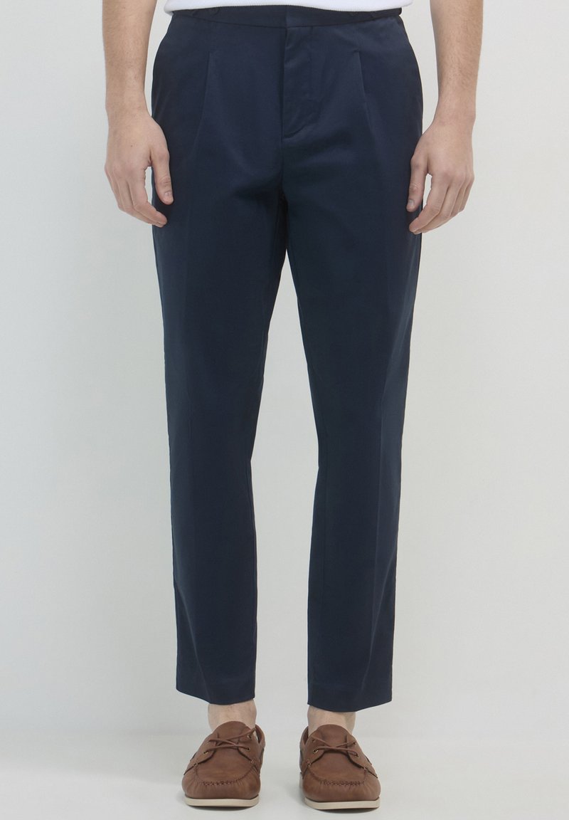 Homme portant un pantalon ajusté bleu marine et des mocassins en cuir marron, se tenant devant un fond clair et unicolore.