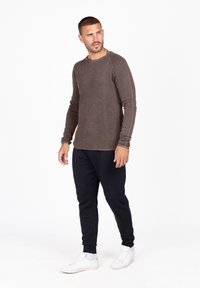 Camicia lunga marrone a coste abbinata a pantaloni jogger blu e sneakers bianche. La camicia presenta un collo rotondo e polsini a costine.