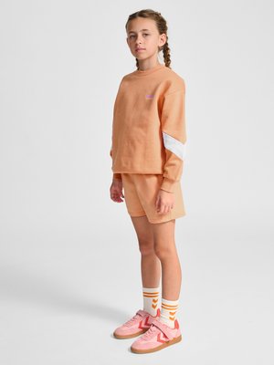 Jong meisje met gevlochten haar, draagt een perzikkleurige trui, bijpassende korte broek, witte sokken met oranje strepen en roze sneakers, staand tegen een effen achtergrond.