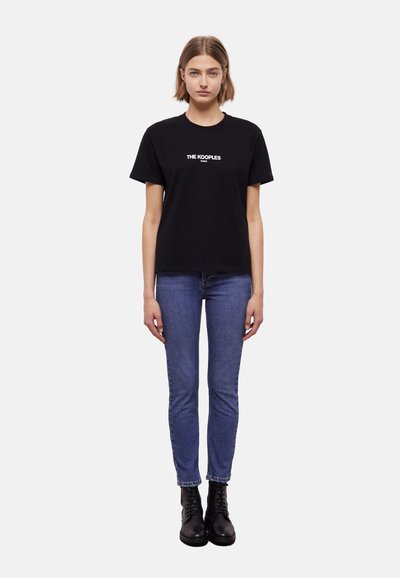 T-shirt noir en coton avec le texte blanc "THE KOOPLES PARIS", associé à un jean skinny bleu et des bottines à lacets noires.