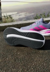 Chaussure de sport gris clair avec des accents roses, dotée d'une semelle en caoutchouc texturée et d'un intercalaire rembourré, posée sur une surface en asphalte.