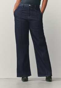 Blugi cu talie înaltă, cu croială largă și culoare albastru închis, având buzunare frontale, cusături vizibile și o textură subtilă a țesăturii.