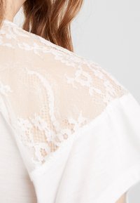 Top blanco con acentos de encaje en los hombros, que presenta una textura suave y un delicado patrón floral. La tela es ligera y semitranslúcida.