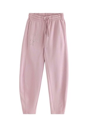 Ung person med krøllet hår iført en lyserød sweatshirt og matchende bukser med stjernedesign, står med hænderne i lommerne og smiler.
