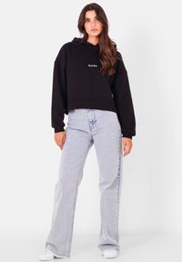 Svart kort hoodie med broderad logotyp på bröstet, matchad med ljusblå högmidjade vida jeans och vita skor.