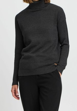 Vrouw draagt een donkergrijze, gestructureerde coltrui met geribbelde manchetten en zoom, gecombineerd met een zwarte broek, tegen een effen achtergrond.