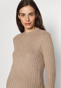 Maglione a coste beige con collo alto e maniche lunghe, progettato per adattarsi comodamente a un pancione. Texture morbida, silhouette aderente.