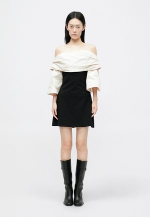 Juoda trumpa suknelė su balta, nuoga pečių apdaila, turinti balioninius rankovės ir juodas aulinukus iki kelių, pristatyta prieš paprastą foną.