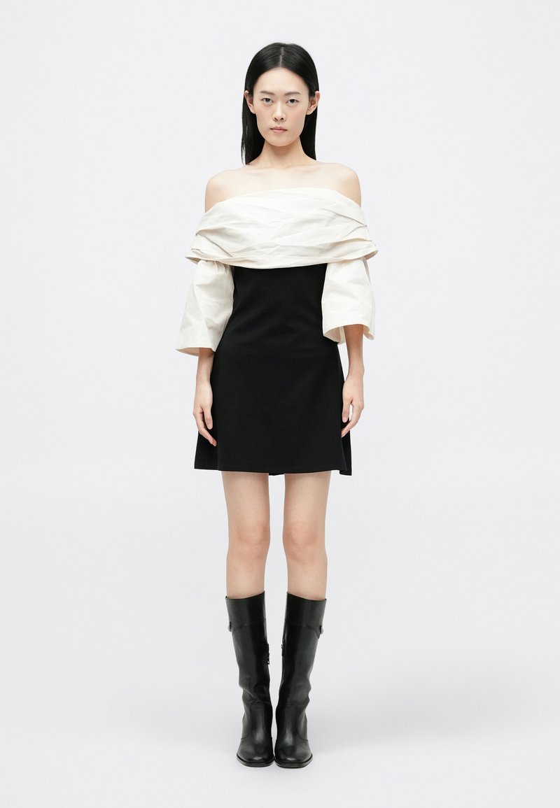 Crna kratka haljina s bijelim, asimetričnim prekrivačem, koji ima balon rukave, i crne čizme do koljena, prikazana na jednostavnoj pozadini.