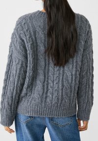 Pull gris en maille avec motifs torsadés, manches larges et ourlet côtelé. Porté sur un jean en denim avec poches arrière visibles.